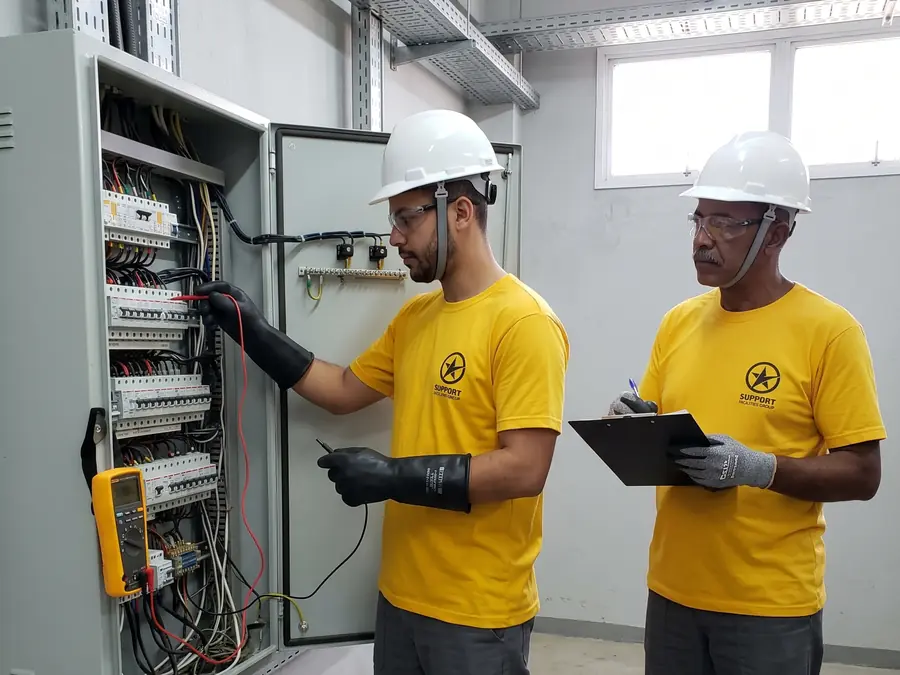 Técnicos com capacete e luvas inspecionando um quadro elétrico